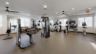 Located at 1500 Cherry St in Placentia, California, this fitness center offers a comprehensive exercise facility designed to support a wide range of workout routines. The space is contained on a single floor and provides a generously sized open layout that accommodates both strength training and cardiovascular equipment without feeling crowded.

The exercise room features durable speckled rubber flooring throughout, ideal for high-impact activity and equipment placement. Overhead, recessed lighting is complemented by sleek ceiling fans that promote air circulation during intense workouts. Multiple windows along the walls invite natural light and provide views of the surrounding exterior, keeping the environment bright and open.

The strength training area is well-appointed with a multi-functional cable machine system, several selectorized resistance machines for targeted muscle groups, a dedicated free weights section with a tiered dumbbell rack, and a weight bench. Large wall-mounted mirrors allow users to monitor their form throughout their sessions.

The cardio section lines one side of the room and includes treadmills, elliptical trainers, and upright stationary bikes, several of which are equipped with individual digital display screens. A motivational "Mamba Mentality" framed print and a neon accent sign add personality to the space. A wall-mounted television and staircase entry area complete this fully functional, modern fitness facility.