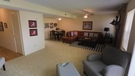 This two-story space features an open-concept main living area that combines a comfortable sitting room with a casual dining space. The living area is furnished with a rich brown sectional sofa accented by decorative pillows, complemented by a bold houndstooth-patterned area rug that adds visual contrast to the light-colored carpeted flooring. A ceiling fan with a light fixture is centrally positioned overhead, providing both airflow and ambient lighting. The seating arrangement is completed with additional upholstered chairs, creating an inviting and functional layout.

Adjacent to the living area, the dining space accommodates a dark wood table with matching chairs, ideal for everyday meals. The walls feature framed artwork and University of Alabama-themed décor, giving the space a distinct personality. Neutral-toned walls and carpeting throughout contribute to a cohesive, warm ambiance. A second ceiling-mounted light fixture illuminates the dining zone. The overall layout offers an efficient use of open floor space, with clear sightlines between the living and dining areas, making the main level feel connected and welcoming.