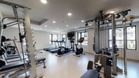 This fitness center is designed to accommodate a comprehensive range of workout routines within a single, open-plan space. The room features a light-toned, durable flooring surface throughout, recessed ceiling lighting, and multiple windows that allow natural light to filter in, creating a bright and energizing environment.

The cardio area is equipped with treadmills, an elliptical trainer, and a stationary bike, all positioned near the windows to allow users to exercise while enjoying outdoor views. A wall-mounted flat-screen television is installed nearby for entertainment during workouts.

A dedicated strength training section features a multi-station cable machine system, offering a wide variety of resistance exercises. Adjacent to this, a free weights area includes a full dumbbell rack with an organized selection of weights, accompanied by adjustable benches and a squat rack for functional strength training.

An accent wall in teal adds a distinct visual element to the strength zone, contrasting with the otherwise neutral white and gray tones of the space. Entry to the fitness center is through glass-paneled double doors, providing visibility into the room. The overall ambiance is clean, modern, and functional, catering to a broad range of fitness needs.