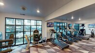 This fitness center is thoughtfully designed to support a comprehensive workout experience across multiple interconnected zones. The space is anchored by a geometric-patterned carpet in neutral tones of brown, gray, and white, which extends throughout and lends a modern, cohesive aesthetic.

The cardio zone is lined with large, black-framed windows that span nearly floor to ceiling, flooding the area with natural light and providing views of the surrounding exterior. Multiple treadmills, ellipticals, a StairMaster, and a rowing machine are arranged in this section, complemented by wall-mounted flat-screen televisions for entertainment during workouts.

Adjacent to the cardio area, a strength training zone houses a cable machine, a dumbbell rack visible through a large mirror wall, and various resistance equipment. The full-length mirrors amplify the sense of depth and allow users to monitor their form effectively.

Near the entrance, a transition area features a dark accent wall with open industrial-style shelving, a built-in cabinet, and a small appliance station—offering a functional space for check-in or equipment storage. Recessed lighting throughout provides consistent, well-distributed illumination, while double doors provide clear access between workout zones.
