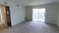 This single-floor space offers a clean and functional layout designed with simplicity and comfort in mind. The living area is the focal point of the space, featuring soft neutral carpeting that extends across the room, providing a warm and understated foundation. Crisp white walls keep the interior feeling bright and open, offering a versatile canvas for a variety of furnishing styles.

A large sliding glass door serves as a standout architectural element, flooding the room with abundant natural light and framing a tranquil view of lush, wooded surroundings. The door provides direct access to an outdoor area, seamlessly connecting the interior with the natural landscape beyond. Vertical blinds accompany the sliding door, offering adjustable light control and privacy.

The space is equipped with practical built-in features, including ceiling ventilation and baseboard heating units that ensure year-round climate comfort. The overall ambiance is calm and neutral, making it well-suited for those who appreciate a straightforward, move-in-ready environment. The wooded backdrop visible through the glass door adds a refreshing natural element that enhances the sense of openness and tranquility throughout the living space.