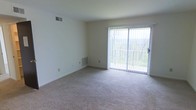 This single-floor space offers a clean and functional layout designed with simplicity and comfort in mind. The living area is the focal point of the space, featuring soft neutral carpeting that extends across the room, providing a warm and understated foundation. Crisp white walls keep the interior feeling bright and open, offering a versatile canvas for a variety of furnishing styles.

A large sliding glass door serves as a standout architectural element, flooding the room with abundant natural light and framing a tranquil view of lush, wooded surroundings. The door provides direct access to an outdoor area, seamlessly connecting the interior with the natural landscape beyond. Vertical blinds accompany the sliding door, offering adjustable light control and privacy.

The space is equipped with practical built-in features, including ceiling ventilation and baseboard heating units that ensure year-round climate comfort. The overall ambiance is calm and neutral, making it well-suited for those who appreciate a straightforward, move-in-ready environment. The wooded backdrop visible through the glass door adds a refreshing natural element that enhances the sense of openness and tranquility throughout the living space.