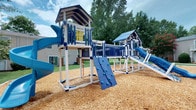 This community amenity space combines an outdoor playground and an indoor fitness center, providing a well-rounded environment for recreation and exercise. The playground features a striking blue-and-white multi-level play structure set over a wood chip safety surface. The structure includes covered upper platforms with pitched roof shelters, blue vertical rail fencing, multiple slides, a rock climbing wall with molded hand and foot holds, and interconnecting bridges — all framed by mature trees and open green lawn. The design encourages active, imaginative play in a safe and visually engaging outdoor setting. The indoor fitness center is bright and purposefully designed, with walls painted in warm yellow accented by bold orange and dark brown horizontal stripes that give the room a motivating, energetic feel. Track lighting illuminates the rubber-floored space, which is equipped with a multi-station cable weight machine, treadmills, an elliptical trainer, a stationary bike, and free weights. Full-length mirrors line one wall, enhancing the sense of space and allowing users to monitor their form. Windows provide natural light and ventilation, while the overall layout supports a comfortable and functional workout experience.