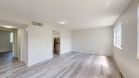 This single-floor unit presents a clean, contemporary aesthetic defined by a neutral white and grey palette and light hardwood-style flooring that runs consistently throughout. The open living area is spacious and airy, benefiting from natural light that filters in through windows fitted with vertical blinds. A flush-mount ceiling light fixture provides additional illumination, while a centrally placed HVAC vent ensures year-round climate control.

The kitchen is well-appointed with crisp white shaker-style cabinetry accented by matte black hardware, light quartz-style countertops, and stainless steel appliances including a Whirlpool range with an overhead ventilation hood and a full-size refrigerator. The layout provides ample counter and storage space.

The bedroom is comfortably sized with a ceiling fan and light kit, a window with vertical blinds offering natural light, and the same light flooring found throughout the unit. An additional room features dual windows framing a pleasant green outdoor view, adding a refreshing natural element to the interior.

The bathroom features a white tiled tub-shower combination, modern matte black fixtures and accessories, and a clean, minimalist design. Overall, the unit delivers a bright, move-in ready living environment with cohesive modern finishes.