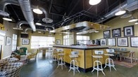 This single-floor space showcases a striking industrial-chic aesthetic that blends raw architectural elements with vibrant design choices. The centerpiece is a large, boldly yellow bar counter with a granite-style top, surrounded by white metal adjustable bar stools that reinforce the space's contemporary yet retro character. A suspended chrome hood and a hanging glass rack above the bar add functional elegance to the design.

The ceiling is finished in matte black, allowing the exposed ductwork, ventilation pipes, and industrial pendant lighting to stand out as intentional design features rather than purely utilitarian elements. Polished concrete floors run throughout, contributing to the open, loft-like feel of the space.

Natural light filters in through several windows fitted with horizontal blinds along one wall, brightening the interior and complementing the warm yellow tones. A lounge area with upholstered seating is visible in the background, offering a more relaxed setting within the same open floor plan. Black-and-white framed photographs on the walls add a curated, urban touch. The overall ambiance is energetic, social, and well-suited for gathering, hosting, or collaborative use.