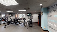 Located at 325 Cordova St in Pasadena, California, this fitness center offers a well-organized, single-floor layout designed to support a comprehensive workout experience. The facility is divided into distinct zones, each dedicated to a specific type of training. The cardio area is equipped with treadmills, elliptical trainers, a recumbent bike, and a stair climber, all positioned near windows that allow natural light to filter in and provide views of the surrounding greenery. Mounted flat-screen televisions are placed throughout for entertainment during workouts. The strength training section features a multi-functional cable machine system, a selectorized weight machine, a captain's chair, and additional specialty equipment, offering a full range of resistance training options. A dedicated free weights area includes a tiered dumbbell rack, adjustable benches, stability balls, and medicine balls, complemented by mirrored walls that enhance the sense of space. The flooring throughout is durable rubber gym matting, suitable for high-traffic use. A teal accent column and a motivational typography wall mural featuring words like "strength," "challenge," and "dedication" add visual character to the space. A wall clock and vending machine provide practical amenities. Recessed and panel lighting ensures consistent illumination across all areas of the facility.