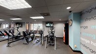 Located at 325 Cordova St in Pasadena, California, this fitness center offers a well-organized, single-floor layout designed to support a comprehensive workout experience. The facility is divided into distinct zones, each dedicated to a specific type of training. The cardio area is equipped with treadmills, elliptical trainers, a recumbent bike, and a stair climber, all positioned near windows that allow natural light to filter in and provide views of the surrounding greenery. Mounted flat-screen televisions are placed throughout for entertainment during workouts. The strength training section features a multi-functional cable machine system, a selectorized weight machine, a captain's chair, and additional specialty equipment, offering a full range of resistance training options. A dedicated free weights area includes a tiered dumbbell rack, adjustable benches, stability balls, and medicine balls, complemented by mirrored walls that enhance the sense of space. The flooring throughout is durable rubber gym matting, suitable for high-traffic use. A teal accent column and a motivational typography wall mural featuring words like "strength," "challenge," and "dedication" add visual character to the space. A wall clock and vending machine provide practical amenities. Recessed and panel lighting ensures consistent illumination across all areas of the facility.