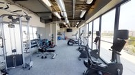 This contemporary fitness center is designed to deliver a complete workout experience in a bright, open environment. The space is lined with expansive floor-to-ceiling windows along one wall, flooding the interior with natural light and offering sweeping views of the surrounding cityscape — creating an energizing atmosphere for exercise.

The layout is thoughtfully organized to maximize usability. Along the windowed side, a row of cardio machines — including treadmills, stationary bikes, and ellipticals — allows members to work out while taking in the outdoor scenery. On the opposite side, a well-stocked free weights area features a full rack of dumbbells in varying sizes, a weight bench, and additional functional training accessories such as medicine balls and stability balls, catering to a wide range of strength training routines.

The industrial-style ceiling, with exposed ductwork and modern overhead lighting, adds an urban aesthetic to the space. Neutral-toned carpeted flooring provides comfort underfoot throughout the workout area. A wall-mounted flat-screen television is positioned for easy viewing from the cardio section, adding an entertainment element to training sessions. Overall, the fitness center combines practical amenities with a clean, modern design to support a variety of fitness goals.