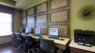 This single-floor business center is designed with both functionality and contemporary style in mind. The space features a row of individual computer workstations arranged along a continuous built-in desk surface that runs the length of one wall, providing ample workspace for multiple users simultaneously. Each station is equipped with a desktop computer and keyboard, and is paired with a sleek, ergonomic black office chair, ensuring comfortable extended use.

The defining architectural feature of the space is the striking accent wall, which is paneled in dark espresso-toned framing and adorned with decorative laser-cut panels displaying an intricate geometric lattice pattern. Subtle horizontal accent lighting is integrated between the panel rows, casting a warm glow that highlights the textural detail of the wall art. The surrounding walls are painted in a bold olive green, adding a vibrant, modern contrast to the dark paneling.

The overall ambiance is polished and professional, with a design aesthetic that balances decorative flair with practical utility. The space is well-suited as a shared business center or computer lab within a hospitality or residential community setting.
