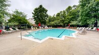 Located in Annapolis, Maryland, this outdoor community amenity area centers around a generously sized swimming pool with clear turquoise water. The pool features a classic rectangular shape with an angled extension, entry steps with stainless steel handrails, and clearly marked depth indicators along its perimeter. The surrounding concrete sun deck provides ample space for relaxation and is furnished with numerous lounge chairs and red patio umbrellas positioned throughout the area.

The pool area is enclosed by a sleek black iron fence, offering both security and an open, airy feel. Mature deciduous trees frame the space on all sides, providing natural shade and a lush, green backdrop that enhances the overall ambiance. Landscape beds filled with low-growing ground cover and ornamental plantings add to the polished, well-maintained aesthetic of the grounds.

Adjacent to the pool, a children's playground is visible behind the fencing, equipped with slides and play structures and surrounded by additional mature trees and manicured landscaping. A brick clubhouse building with traditional architectural details sits nearby, complementing the community's residential character. Additional amenities include outdoor grills and conveniently placed waste receptacles. The entire space is thoughtfully designed to offer a resort-like outdoor experience within a residential community setting.