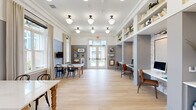 Located at 7100 Summerside Dr in Whitsett, NC, this thoughtfully designed community room blends dining and productivity into a single, cohesive space. Light hardwood flooring runs throughout, complementing the crisp white crown molding and two-tone gray and white walls that give the room a polished, modern character. A row of globe pendant lights in matte black fixtures hang from the ceiling, providing warm, even illumination across the entire area. The dining section features sturdy farmhouse-style tables with white marble tops and turned wood legs, paired with classic black bentwood chairs, while wall-mounted flat-screen televisions and a curated gallery wall of framed artwork add visual interest. Tall windows dressed with floor-length linen drapes invite natural light and offer views of the surrounding outdoor community. A dedicated co-working area lines one side of the room, offering built-in desk stations set against a textured whitewashed brick accent wall, each furnished with leather chairs and computer monitors. Above, expansive open shelving displays books, plants, and decorative objects. A beverage station with blue-gray cabinetry, brass hardware, and a coffee machine anchors the workspace side of the room. Glass double doors provide seamless access to the outdoors, enhancing the bright and welcoming ambiance throughout.
