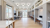 Located at 7100 Summerside Dr in Whitsett, NC, this thoughtfully designed community room blends dining and productivity into a single, cohesive space. Light hardwood flooring runs throughout, complementing the crisp white crown molding and two-tone gray and white walls that give the room a polished, modern character. A row of globe pendant lights in matte black fixtures hang from the ceiling, providing warm, even illumination across the entire area. The dining section features sturdy farmhouse-style tables with white marble tops and turned wood legs, paired with classic black bentwood chairs, while wall-mounted flat-screen televisions and a curated gallery wall of framed artwork add visual interest. Tall windows dressed with floor-length linen drapes invite natural light and offer views of the surrounding outdoor community. A dedicated co-working area lines one side of the room, offering built-in desk stations set against a textured whitewashed brick accent wall, each furnished with leather chairs and computer monitors. Above, expansive open shelving displays books, plants, and decorative objects. A beverage station with blue-gray cabinetry, brass hardware, and a coffee machine anchors the workspace side of the room. Glass double doors provide seamless access to the outdoors, enhancing the bright and welcoming ambiance throughout.