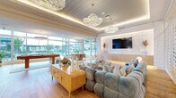 Located at 3615 Tryclan Drive in Charlotte, NC, this sophisticated amenity lounge spans a generous single floor and showcases a carefully curated blend of modern and coastal-inspired design. The open-concept layout flows seamlessly across wide-plank hardwood flooring, connecting distinct functional zones within one cohesive space.

The main lounge area serves as the centerpiece, featuring plush tufted sectional sofas in soft blue velvet arranged around a sculptural glass-top coffee table. Statement cloud-inspired glass chandeliers hang from tray ceilings with integrated ambient lighting, creating a warm, layered glow throughout. An elegant decorative wall panel with undulating wave patterns adds artistic depth to this seating zone.

A dedicated entertainment area includes a wall-mounted flat-screen TV set against a backlit built-in panel, while a billiards table anchors a secondary lounge zone bathed in natural light. Floor-to-ceiling windows along the perimeter provide sweeping outdoor views and flood the interior with abundant daylight.

The wet bar area is equally refined, featuring a dramatic marble-topped island, a textured geometric tile accent wall, stainless steel appliances, and a distinctive herringbone wood ceiling detail overhead. A separate café-style seating nook with banquette seating and round tables sits adjacent to full-height glass entry doors, offering a bright, welcoming gathering spot.