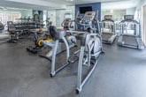 The Bloomfield Square Fitness Center at 3161 Bloomfield Ln in Auburn Hills, Michigan, is a comprehensively outfitted, single-floor facility designed to support a wide range of workout routines. The space features rubber flooring throughout, providing a durable and safe surface suited for both cardio and strength training activities.

The cardio zone is well-stocked with multiple treadmills, elliptical machines, and stationary bikes, including an air resistance bike. Large mirrors line the walls, visually expanding the space and allowing users to monitor their form. Natural light filters in through window treatments on one side, complemented by recessed ceiling lighting and a ceiling fan that keeps the environment comfortable during workouts.

The strength training area includes a multi-station cable machine, adjustable weight benches, and a full rack of dumbbells ranging across various weights, providing ample options for resistance training. Wall-mounted equipment and additional free weights round out the strength offerings.

Near the entrance, a dedicated utility area features a water cooler, a rack of yoga and exercise mats in multiple colors, and posted Bloomfield Square Fitness Center rules. Motivational wall art adds a welcoming touch to the space. An adjacent room accessible through a white paneled door offers additional storage or amenity space. The overall ambiance is clean, functional, and well-maintained, making it a practical fitness resource for residents.