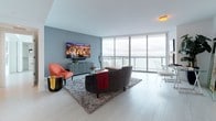 Unit 49A is a thoughtfully designed single-floor residence that combines modern aesthetics with high-end functionality. The open-concept living area is anchored by dramatic floor-to-ceiling windows that frame sweeping panoramic views of the skyline and waterfront, flooding the space with natural light. Light wood flooring flows throughout, complementing the contemporary color palette of soft grays and crisp whites.

The kitchen is outfitted with sleek, high-gloss white cabinetry, dark countertops, under-cabinet LED accent lighting, and premium stainless steel Bosch appliances including a built-in oven, microwave, and French door refrigerator. A central island with bar seating provides additional workspace and casual dining options.

The primary bedroom features direct access to expansive views through floor-to-ceiling windows, a tufted upholstered headboard, and a walk-in closet. A second bedroom is equally well-appointed with its own closet and wall-mounted television. Both bathrooms are finished with striking marble-style tile on floors and walls, glass-enclosed showers, soaking tubs, white vanities, and illuminated mirrors, delivering a clean, spa-like atmosphere throughout.