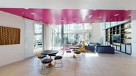 Located in the heart of Downtown Los Angeles's Arts District, this single-floor community amenity space at 950 E Third St offers a striking blend of contemporary design and bold artistic expression. The centerpiece of the space is a generously proportioned lounge area distinguished by a vivid, glossy magenta ceiling that contrasts dramatically with the clean white walls and large-format neutral floor tiles beneath. Floor-to-ceiling glass walls and sliding doors flood the interior with abundant natural light while offering unobstructed views of an adjacent outdoor courtyard and the surrounding residential architecture.

The lounge is thoughtfully furnished with eclectic, design-forward pieces—gold wire-frame chairs paired with dark round bistro tables, plush navy sofas, and playful accent seating in matching pink upholstery. A sculptural gold animal figurine serves as a conversation-starting art piece, while curated wall art, including a large abstract pink canvas and a geometric textile work, reinforce the space's gallery-like atmosphere.

A separate, fully equipped communal kitchen runs parallel to the lounge, featuring sleek white cabinetry, dark stone countertops, a gas cooktop island with a stainless steel range hood, a mosaic tile backsplash, and stainless steel appliances. A transitional corridor with direct exterior access connects the kitchen to the lounge, completing this dynamic and inviting communal environment.