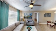 This single-story, two-bedroom, two-bathroom residence offers a thoughtfully organized layout across a comfortable footprint. The living room serves as the heart of the home, featuring light wood-style flooring, a ceiling fan with integrated lighting, and a textured ceiling. Large windows dressed with teal grommet curtains invite natural light while adding a pop of color against the warm neutral walls. A glass-top coffee table and tufted sofa contribute to the room's modern, casual aesthetic.

The galley-style kitchen is efficiently designed with white cabinetry, granite-look countertops, and white appliances, including a range and full-size refrigerator. The adjacent dining area is highlighted by a contemporary drum pendant light and provides a dedicated space for seated meals.

Both bedrooms are carpeted and include ceiling fans, windows with blinds, and generous closet space. One bedroom features an en-suite bathroom, while the second bathroom offers a white vanity with a large mirror, overhead storage cabinet, and a shower-tub combination. The neutral color palette and consistent finishes throughout create a cohesive, comfortable atmosphere.