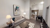 This well-appointed single-floor residence showcases a thoughtfully designed open-concept layout that maximizes both space and functionality. The living area is anchored by a sleek, light-toned sofa accented with decorative pillows, complemented by a patterned area rug and a minimalist coffee table. Towering ceilings create an impressive sense of volume and airiness throughout the space, while track lighting adds a modern, gallery-like quality to the room. A large framed abstract artwork adorns the clean white walls, lending the space a sophisticated, curated feel. Wide-plank hardwood flooring in a warm grey tone runs continuously throughout, tying the rooms together in a cohesive aesthetic. The kitchen is outfitted with contemporary wood-toned cabinetry, stainless steel appliances, and a functional breakfast bar, making it equally suited for casual dining and meal preparation. Adjacent to the kitchen, a compact dining area with a dark table and chairs provides a defined space for meals. A louvered door adds a classic architectural detail and offers practical storage access. Overall, the space exudes a clean, modern ambiance with a neutral palette, elevated finishes, and an efficient, livable design.