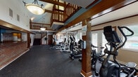 Located at 940 Spring Forest Rd in Greenville, NC, this two-story community amenity building combines fitness, social, and relaxation spaces under one roof. The ground floor is anchored by a spacious fitness center with durable rubber flooring, housing a comprehensive array of equipment including weight machines, ellipticals, stationary bikes, and treadmills. Soaring vaulted ceilings with exposed wood beams and structural columns lend the gym a distinctive architectural character, while multiple sets of French doors and large windows invite abundant natural light. A prominent brick-faced bar counter with rich wood cabinetry and built-in shelving serves as a social focal point on the main level, alongside access to a kitchen area. The second floor offers a welcoming lounge area with hardwood-style flooring and a vaulted ceiling, furnished with comfortable seating. An adjacent dining area features a high-top table with seating for four, framed art, and sliding glass doors leading to an outdoor balcony. A large open room on this level showcases a dramatic bank of oversized windows overlooking the outdoor pool and surrounding greenery, creating a bright and airy atmosphere. A wood-railed staircase connects both levels, tying together the warm, craftsman-inspired aesthetic found throughout the building.