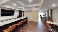 This thoughtfully designed single-floor space showcases a seamless open-concept layout that connects the kitchen, living, bar, and dining areas into one cohesive and elegant environment. The gourmet kitchen is appointed with dramatic two-tone cabinetry — dark lower cabinets paired with white upper cabinets featuring glass-front doors — and striking veined granite countertops. A full suite of stainless steel appliances, including a gas range with a statement hood vent, dishwasher, and French door refrigerator, elevates the kitchen's functionality. A subway tile backsplash and geometric brass pendant lights add refined finishing touches. Rich hardwood flooring flows throughout the entire space, grounding the modern aesthetic. The living area is anchored by a textured stacked-stone accent wall housing a sleek linear fireplace and a wall-mounted television, beneath an impressive coffered ceiling that adds architectural depth. Adjacent to the living space, a sophisticated wet bar features a long marble-topped counter with leather bar stools, set against a dark feature wall. The dining area benefits from large windows and a glass door that invite natural light and offer views of an outdoor area. Decorative columns subtly define zones while maintaining the open flow of the space.