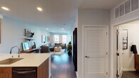 This well-appointed single-floor residence showcases a thoughtfully designed open-concept layout that seamlessly connects the kitchen, dining, and living areas. Dark hardwood flooring runs throughout, providing a warm, cohesive foundation that complements the clean white walls and modern finishes.

The kitchen is a standout feature, outfitted with rich walnut cabinetry, white quartz countertops, and black stainless steel appliances including a side-by-side refrigerator, range, and built-in microwave. A spacious island with bar seating offers an ideal spot for casual dining, while under-cabinet lighting adds a refined touch.

The open living area is bathed in natural light courtesy of large windows with sheer curtains, offering views of the surrounding urban landscape. Recessed lighting throughout enhances the bright, airy ambiance. A louvered closet door adds a classic architectural detail near the entryway.

The bedroom is a serene retreat, featuring a bold green accent wall, large windows, and dark hardwood floors that continue from the main living space, creating a calming and cohesive environment.

The bathroom is finished with a walnut vanity, white quartz countertop, a large mirror, and dark hardware, maintaining the consistent warm-toned, modern aesthetic found throughout the residence.