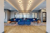 Located at 800 Marietta St NW in Atlanta's vibrant Westside neighborhood, this single-floor living space offers a generously sized, open-plan layout designed for both comfort and social interaction. The main living area is anchored by a large, bold blue sectional sofa arranged over a colorful area rug, flanked by round wood-and-metal coffee tables and leather pouf ottomans, creating an inviting and eclectic atmosphere. Warm hardwood-style flooring runs throughout, complementing the cool gray walls and contemporary aesthetic. A grid of elegant globe pendant lights illuminates the central lounge area, while floor-to-ceiling steel-framed windows and glass doors flood the space with natural light and provide views to an outdoor terrace. Structural columns with dark metal accents add an industrial character to the open layout. A built-in cabinetry station with countertop and integrated appliances serves as a practical hub within the space. A cozy nook features vibrant yellow banquette seating with patterned upholstery, a sculptural gold pendant light, and a window overlooking the city. A media wall with a large flat-screen TV and stone accent wall anchors an additional lounge zone. The overall ambiance is modern, colorful, and thoughtfully curated, making this space well-suited for community gathering and relaxation.