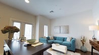 This studio-style unit presents a thoughtfully organized single-floor layout that maximizes functionality within a compact footprint. The open-concept design seamlessly connects the living and dining areas, creating a cohesive and practical living environment. The living space features smooth white walls accented by recessed lighting and ceiling vents, contributing to a clean, contemporary aesthetic. Natural light filters into the room through multiple windows fitted with horizontal blinds, brightening the interior and creating an inviting atmosphere. The dining area is anchored by a dark-toned table, providing a defined space for meals while remaining visually integrated with the rest of the unit. A glass-paneled door offers additional natural light and potential outdoor access. The living area provides ample room for comfortable seating and décor, as illustrated by the generous sectional sofa arrangement shown. Neutral wall tones throughout the unit serve as a versatile backdrop, suitable for a variety of interior styles. The overall ambiance is modern and understated, with an efficient use of space that suits individuals seeking a streamlined, low-maintenance living environment.