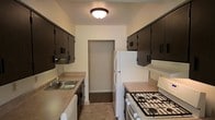 This two-story residence offers a practical and well-organized layout designed for efficient everyday living. The kitchen follows a classic galley configuration, with parallel countertops running along both sides to maximize workspace and workflow. Dark espresso-toned cabinetry lines the upper walls on both sides, providing ample storage while creating a strong visual contrast against the light-colored walls and tile flooring. The countertops feature a neutral laminate finish, complementing the overall color palette. White appliances, including a gas range and a full-size refrigerator, are neatly integrated into the layout, keeping the space cohesive and clean. A stainless steel sink is positioned along one counter, accompanied by standard under-cabinet storage below. A flush-mount ceiling light provides consistent overhead illumination throughout the kitchen. At the far end of the kitchen, a doorway opens into an adjacent room, suggesting a connected and accessible floor plan. The two-story layout of the residence allows for a natural separation of living and private spaces across floors, contributing to an organized and functional home environment.