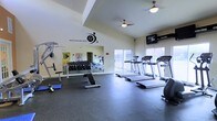 This health and wellness fitness center is designed to support a comprehensive workout experience within a single, open-plan space. The room features vaulted ceilings with a ceiling fan that promotes airflow and comfort during exercise. Large windows along one wall flood the interior with natural light and provide views of the outdoors, creating an energizing atmosphere. A wall-mounted flat-screen TV adds entertainment options for those on cardio equipment. The flooring is covered with durable rubber gym matting, ideal for both safety and performance. A row of treadmills lines one side of the room, catering to cardio enthusiasts, while a dedicated free weights area includes a full rack of dumbbells and a workout bench for strength training. A multi-use cable machine provides additional resistance training options. Floor-to-ceiling mirrors expand the sense of space while allowing users to monitor their form. The "Health and Wellness" logo displayed prominently on the wall reinforces the space's dedicated fitness purpose. Overall, the facility strikes a balance between functionality and comfort, offering a well-rounded environment for residents or members to pursue their fitness goals.