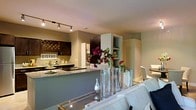 This well-appointed single-floor residence offers a thoughtfully designed open-concept layout that connects the living and kitchen areas in a cohesive, flowing arrangement. The kitchen is a standout feature, showcasing dark cabinetry with a rich wood finish, granite countertops, and a peninsula that doubles as a breakfast bar — ideal for casual dining or entertaining. A decorative wine rack mounted on the wall adds a charming bistro-inspired character, complemented by track lighting overhead that provides warm, focused illumination throughout the space. Built-in shelving offers both functional storage and display opportunities. The living area transitions seamlessly from the kitchen, anchored by a plush white sofa and a mirrored coffee table that reflects light and adds a sense of openness. Decorative glass vases with vibrant floral arrangements bring a lively pop of color to the space. The neutral, earthy wall tones throughout create a calm and inviting ambiance, while the overall design balances modern aesthetics with warm, personalized touches. The single-level layout ensures easy accessibility and a comfortable flow between spaces, making it well-suited for both everyday living and hosting guests.