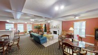 This single-floor community space exudes warmth and comfort through its thoughtful design and inviting atmosphere. The main lounge area is anchored by a classic white-framed fireplace with a decorative mantel, serving as a natural focal point for the room. Walls painted in a rich terracotta hue create a warm, welcoming ambiance that complements the carefully curated furnishings throughout.

The seating area features a plush teal sofa paired with coordinating armchairs, arranged around a distinctive yellow upholstered coffee table atop a patterned area rug — ideal for casual conversation or relaxation. A separate dining arrangement with a glass-top table and traditional wooden chairs offers additional functionality for group gatherings or activities.

Architectural details include coffered ceilings adorned with elegant crown molding and a textured finish, adding visual depth and character to the space. Track lighting and ceiling-mounted fixtures ensure the room is well-lit throughout. Large windows along the perimeter allow natural light to filter in, enhancing the overall brightness of the interior. Lush indoor plants are thoughtfully placed to bring a touch of nature indoors, further enriching the space's warm and lively character.
