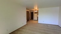 This single-floor residential unit offers a straightforward, functional layout designed for comfortable everyday living. The main living area is finished with wall-to-wall carpeting and neutral-toned walls, creating a clean and versatile space ready for furnishing. A large sliding glass door dominates one wall of the living room, flooding the interior with natural light and providing direct access to a private outdoor deck — an ideal spot for relaxing or enjoying the surrounding views of the neighboring brick building and open sky.

Transitioning toward the back of the unit, the open layout connects the living area to the entryway and kitchen zone. The entry features dark-toned wood doors, adding a warm contrast to the light-colored walls. The kitchen is finished with tile flooring and is equipped with a refrigerator, cabinetry, and an overhead ceiling fan with lighting, providing functional illumination for both the kitchen and adjacent dining or living space.

Throughout the unit, textured ceilings and dark wood trim accents on the doors and window frames add subtle character. The overall ambiance is bright, neutral, and move-in ready, offering a practical living environment with cohesive finishes throughout its single-level floor plan.
