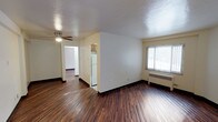 This single-floor space offers a clean and functional layout designed for comfortable everyday living. The main living area is finished with rich, dark hardwood-style flooring that runs throughout, creating a warm and cohesive aesthetic. A ceiling fan is centrally mounted, providing both lighting and air circulation. A large window fitted with vertical blinds allows natural light to fill the space while offering a view of the outdoors. A baseboard heating unit beneath the window ensures year-round comfort. The textured ceiling adds a subtle architectural detail to the otherwise clean, neutral interior. The living area flows naturally into a partially open kitchen, visible through a dividing half-wall. The kitchen features light-toned cabinetry and a white refrigerator, contributing to a bright and practical cooking space. A hallway leads further into the unit, connecting to additional rooms that continue the same hardwood-style flooring. The walls throughout are finished in a soft, neutral tone that complements the darker flooring. Overall, the space presents a straightforward and well-maintained interior that balances functionality with a welcoming ambiance.