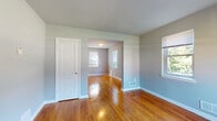 This two-floor home presents a well-maintained and cohesive interior defined by warm hardwood flooring that runs throughout the main living areas and bedrooms, reflecting natural light and adding a timeless character to the space. The main level welcomes you through a front entry with a staircase leading to the upper floor, opening into a bright living area with large windows that overlook the street, filling the space with abundant daylight. The connected layout flows seamlessly into additional rooms, each featuring the same polished hardwood floors, soft gray-toned walls, and white trim detailing. The kitchen is equipped with warm wood cabinetry extending to the ceiling, a granite-style countertop with a stainless steel sink, and a gas range, offering a practical and functional cooking space. The bathroom features classic white subway tile wainscoting, a soaking tub with shower, a toilet, and a vanity sink, with a window providing natural ventilation and light. The upper level hosts several generously sized rooms, each with multiple windows framing views of surrounding greenery. Throughout the home, floor vents, white panel doors, and consistent neutral finishes contribute to a clean, understated aesthetic that is both functional and inviting.