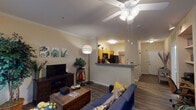 This single-floor residence offers a thoughtfully designed layout that blends comfort and functionality throughout. The open-concept living area is anchored by warm wood-look flooring, crown molding, and a ceiling fan with integrated lighting. A large window dressed with sheer curtains and horizontal blinds allows natural light to fill the space, creating a bright and welcoming atmosphere. The living room flows seamlessly into the kitchen, which is separated by a peninsula counter with granite-style surfaces and a built-in sink — ideal for casual dining or food preparation. The kitchen is equipped with honey-toned cabinetry, black appliances including a range and built-in microwave, and ample counter and storage space. The bedroom is a versatile retreat, featuring crown molding, a ceiling fan, and a built-in desk nook with open shelving — perfect for a home office setup. A sliding glass door in the bedroom provides access to an outdoor area and brings in additional natural light. The bathroom is clean and functional, offering wood-look flooring, a shower-tub combination, and neutral wall tones. Throughout the residence, consistent warm beige tones and crown molding details lend a cohesive and polished character to the space.