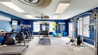 Located at 33451 Mackenzie Way in Lewes, Delaware, this single-floor fitness center is designed to inspire and support a comprehensive workout experience. The space is immediately striking thanks to its bold royal blue walls, which are adorned with motivational phrases such as "Push Yourself Beyond," "Work For It," and "Determination," setting an energetic and focused atmosphere throughout.

The facility is well-lit by a combination of large flush-mount LED panel lights and natural light streaming through multiple windows fitted with white shutters and blinds. A stylish ceiling fan adds airflow and comfort during workouts.

The cardio zone features multiple treadmills and an elliptical trainer, accompanied by a wall-mounted flat-screen TV for entertainment. Adjacent to this area, a dedicated strength training section houses a full rack of dumbbells, a weight bench with rubber floor mat, and a multi-functional cable machine system suitable for a wide range of resistance exercises.

A functional training corner rounds out the equipment offering, with medicine balls, stability balls, resistance bands, and foam rollers neatly organized on dedicated storage racks. Neutral-toned carpet tiles cover the floors throughout, providing a clean and practical surface underfoot. The layout is open and well-organized, allowing for efficient movement between workout zones.