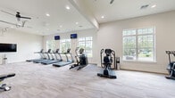 This fitness center offers a spacious, thoughtfully designed workout environment spread across a single floor. The layout is open and expansive, providing ample room for movement between equipment zones. Neutral-toned walls and light-colored carpet flooring give the space a clean, modern aesthetic, while recessed lighting and ceiling fans ensure a comfortable atmosphere throughout.

The main cardio area is lined with a row of treadmills and elliptical machines positioned along a wall of large, multi-pane windows that flood the space with natural light and offer pleasant views of the surrounding greenery and pool area. Wall-mounted flat-screen TVs are positioned above the cardio equipment for entertainment during workouts.

A dedicated strength and functional training zone features a weight bench, air bike, cable machine, and a variety of free weights and resistance tools, including medicine balls and stability balls neatly organized on a rack. A floor-to-ceiling glass partition wall opens toward an outdoor pool area, creating a seamless indoor-outdoor connection.

A separate alcove provides a quieter workout nook with additional equipment, including a recumbent bike and flat bench, topped with its own ceiling fan. The overall ambiance is bright, airy, and professionally appointed, making this fitness center a comprehensive facility suited for a wide range of exercise routines.