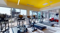 This single-level fitness center makes a striking visual impression through its bold design and well-equipped layout. The most distinctive architectural feature is the vibrant yellow ceiling, which spans the entire space and creates an energetic, motivating atmosphere complemented by recessed linear lighting throughout.

The cardio zone is positioned along the perimeter wall, where an expansive run of floor-to-ceiling glass windows and sliding glass doors floods the area with abundant natural light. This zone is equipped with multiple treadmills, elliptical trainers, and stationary cycling bikes, each featuring individual digital display screens. The large windows offer direct views of an outdoor pool and surrounding amenity areas, creating an open, connected feel during workouts.

The strength training area occupies the interior portion of the space, featuring a comprehensive cable and pulley machine system, a dedicated free weights section with dumbbells, stability and medicine balls stored on wall-mounted racks, and a leg extension bench. Full-length mirrors line the walls of this zone, visually expanding the space and allowing users to monitor their form.

The entrance area features a welcoming welcome mat and a large floor-to-ceiling window overlooking the parking and outdoor areas. Durable, light-toned rubber flooring runs throughout, providing both safety and comfort underfoot.
