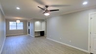 This single-floor space showcases a thoughtfully designed interior with a neutral, modern aesthetic throughout. The main living area is finished with light hardwood-style flooring that brightens the space and complements the soft gray walls, creating a calm and welcoming ambiance. Crown molding adds a refined architectural detail, while crisp white baseboards provide a clean contrast against the flooring.

A ceiling fan with an integrated light fixture is centrally positioned overhead, ensuring both comfort and adequate illumination across the open area. Natural light filters in through a window, enhancing the airy feel of the room.

A standout built-in feature is a recessed desk nook along one wall, complete with a shelf above and a small set of drawers below — a practical and space-conscious design element ideal for a home office setup or study area. The nook is subtly lit, drawing attention to this functional corner.

The overall layout offers an open, uncluttered feel with generous wall space that allows for flexible furniture arrangement. The consistent use of neutral tones, modern fixtures, and clean architectural lines throughout gives the space a cohesive, move-in-ready appeal suited for a variety of lifestyles.