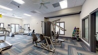 This fitness center offers a thoughtfully designed, single-floor workout space equipped to support a wide range of exercise routines. The interior features neutral-toned walls, a drop-panel ceiling with recessed lighting and ceiling fans for ventilation, and stylish multi-toned blue and grey carpet tiles that add a contemporary feel throughout.

The cardio zone is prominently positioned along a windowed wall, where large framed windows with horizontal blinds allow natural light to fill the space and offer outdoor views. This area is home to treadmills, an elliptical trainer, and a stair climber, each equipped with individual display monitors.

The strength training section features a multi-function cable machine suitable for a variety of resistance exercises, complemented by a dedicated free weight station with a full rack of dumbbells in graduated sizes. A medicine ball rack stocked with color-coded balls of varying weights provides additional functional training options. A rowing machine and adjustable workout bench round out the available equipment.

The layout is open and well-organized, providing ample space to move between stations comfortably. Glass-paneled interior doors connect to an adjoining area, suggesting additional amenities nearby. The overall ambiance is clean, modern, and functional, making it a capable facility for both cardio and strength-focused training.