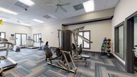 This fitness center offers a thoughtfully designed, single-floor workout space equipped to support a wide range of exercise routines. The interior features neutral-toned walls, a drop-panel ceiling with recessed lighting and ceiling fans for ventilation, and stylish multi-toned blue and grey carpet tiles that add a contemporary feel throughout.

The cardio zone is prominently positioned along a windowed wall, where large framed windows with horizontal blinds allow natural light to fill the space and offer outdoor views. This area is home to treadmills, an elliptical trainer, and a stair climber, each equipped with individual display monitors.

The strength training section features a multi-function cable machine suitable for a variety of resistance exercises, complemented by a dedicated free weight station with a full rack of dumbbells in graduated sizes. A medicine ball rack stocked with color-coded balls of varying weights provides additional functional training options. A rowing machine and adjustable workout bench round out the available equipment.

The layout is open and well-organized, providing ample space to move between stations comfortably. Glass-paneled interior doors connect to an adjoining area, suggesting additional amenities nearby. The overall ambiance is clean, modern, and functional, making it a capable facility for both cardio and strength-focused training.