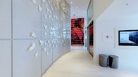 This single-floor space makes an immediate impression through its thoughtfully designed corridor, where art and architecture converge seamlessly. The most prominent feature is a curved, glossy panel wall adorned with dozens of white three-dimensional butterfly sculptures that appear to take flight across its surface, creating a dynamic and visually captivating focal point. The gentle curve of this wall guides visitors naturally through the space, adding an organic flow to the otherwise clean, contemporary lines of the interior. Light-toned large-format floor tiles run the length of the corridor, reflecting the recessed ceiling lighting and contributing to the space's bright, airy atmosphere. At the end of the hallway, a striking large-scale red artwork commands attention, featuring handwritten script text that provides a bold contrast to the predominantly white and grey palette. Floor-to-ceiling glass panels flank the corridor, allowing visibility into adjacent areas and enhancing the sense of openness. The overall ambiance is polished and gallery-like, balancing minimalist architectural elements with carefully selected artistic installations that give the space a distinctive, sophisticated character.