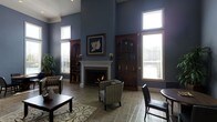 This single-floor space makes a striking impression with its dramatic ceiling height, which amplifies the sense of openness and grandeur throughout the main living area. The walls are finished in a deep blue-gray tone that lends a refined, sophisticated character to the interior. Tall windows with white blinds line the walls, allowing abundant natural light to pour in and brighten the space while framing views of the surrounding exterior.

At the heart of the room sits a classic white-mantled fireplace, serving as a natural focal point and adding warmth and elegance to the space. Flanking the fireplace are two rich, dark wood built-in arched cabinetry units, providing both decorative display space and architectural symmetry. A framed floral artwork is prominently displayed above the mantel, adding an artistic touch.

The flooring consists of neutral stone-tiled surfaces complemented by a decorative area rug, grounding the seating arrangement. Furnishings include patterned accent chairs and a dark wood coffee table, contributing to the space's polished, traditional aesthetic. A dining area with dark leather chairs is visible nearby, adding functional versatility. Lush greenery in the corner introduces a natural element, completing this well-appointed and inviting space.
