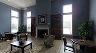 This single-floor space makes a striking impression with its dramatic ceiling height, which amplifies the sense of openness and grandeur throughout the main living area. The walls are finished in a deep blue-gray tone that lends a refined, sophisticated character to the interior. Tall windows with white blinds line the walls, allowing abundant natural light to pour in and brighten the space while framing views of the surrounding exterior.

At the heart of the room sits a classic white-mantled fireplace, serving as a natural focal point and adding warmth and elegance to the space. Flanking the fireplace are two rich, dark wood built-in arched cabinetry units, providing both decorative display space and architectural symmetry. A framed floral artwork is prominently displayed above the mantel, adding an artistic touch.

The flooring consists of neutral stone-tiled surfaces complemented by a decorative area rug, grounding the seating arrangement. Furnishings include patterned accent chairs and a dark wood coffee table, contributing to the space's polished, traditional aesthetic. A dining area with dark leather chairs is visible nearby, adding functional versatility. Lush greenery in the corner introduces a natural element, completing this well-appointed and inviting space.