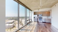 This contemporary high-rise unit offers a striking blend of industrial and modern design, defined by its signature exposed concrete ceilings and expansive floor-to-ceiling glass windows that flood every room with abundant natural light. The open-concept living and kitchen area is anchored by wide-plank hardwood flooring and panoramic city views that stretch across the urban skyline, creating an impressive sense of openness and scale. The kitchen is well-appointed with light wood cabinetry, a white quartz countertop, a hexagonal tile backsplash, and a full suite of stainless steel appliances including a French door refrigerator, electric range, over-the-range microwave, and dishwasher. The bedrooms feature plush carpeting, generous closet space with built-in shelving and hanging rods, and access to the same sweeping city views through large glass windows. The bathrooms are finished with marble-style tile, including a glass-enclosed walk-in shower and a separate tub and shower combination. A dedicated vanity area with a wood-toned cabinet and framed mirror adds a refined touch. Throughout, the interplay of raw concrete, clean white walls, and warm wood tones creates a sophisticated urban aesthetic.