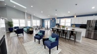 This inviting community room is thoughtfully designed to serve as a versatile gathering space. The open floor plan is bathed in natural light from large, evenly spaced windows along multiple walls, creating an airy and welcoming atmosphere. Recessed lighting and pendant fixtures overhead provide additional illumination throughout the space.

The room is divided into distinct functional zones while maintaining a seamless, open feel. A comfortable lounge area is anchored by a patterned area rug and features navy upholstered armchairs accented with teal throw pillows, paired alongside a teal sofa — all unified by a coordinated, modern color scheme. A casual dining area with round tables and industrial-style seating offers a space for informal meals or small group gatherings.

Along one side of the room, a full kitchen bar takes center stage, complete with white cabinetry, marble-style countertops, a sink, and counter seating with metal bar stools. A coffee station adds a practical touch for everyday use. The kitchen's clean, white-and-blue design complements the bold navy accent wall behind it.

Durable wood-look flooring runs throughout the space, contributing to the room's clean, contemporary aesthetic. Large windows and glass-paned doors provide views to the exterior, enhancing the sense of openness in this well-appointed community gathering area.