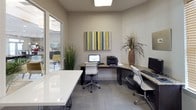 This single-story business center is thoughtfully designed to accommodate multiple users simultaneously in a clean, professional environment. The space features several granite-topped workstations paired with dark wood desk bases and ergonomic mesh chairs, providing both style and comfort for extended use. Carpeted flooring in a neutral gray tone runs throughout, complementing the light gray walls and contributing to a calm, focused atmosphere.

Workstations are equipped with computers, a multifunction printer, and task lighting, ensuring all essential office needs are met. A large skylight and recessed ceiling lights provide consistent, even illumination throughout the space. Decorative touches, including colorful wall art, potted plants, and woven accents, soften the professional environment without distracting from its function.

Large floor-to-ceiling glass windows and a sliding glass door connect the business center to an adjacent lounge area, which is visible through the glazing and features seating, a billiard table, and a television. This transparent boundary allows natural light to filter into the workspace while maintaining a sense of openness. A secondary private workstation nook with a lamp and granite desk offers a quieter, more secluded option for focused individual work.