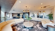 This single-floor space offers a generously sized, open-concept layout that seamlessly blends a sophisticated living area with a fully equipped kitchen. The living room is anchored by a sweeping curved white sectional sofa arranged around an abstract-patterned carpet, complemented by sculptural pendant chandeliers that add a striking focal point overhead. A curated gallery wall of black-and-white framed artwork lends a creative, editorial character to the space. Greenery and potted plants are thoughtfully placed throughout, adding warmth and life to the contemporary palette of dark navy, warm wood, and crisp white tones. Floor-to-ceiling windows along one wall flood the space with abundant natural light and provide open views to the surrounding streetscape, framed by textured stone-clad columns that add an architectural dimension. The kitchen area is equally refined, featuring a large central island with a white countertop, light wood cabinetry, vertically laid tile backsplash, and a live-edge wood dining table with modern seating. A wet bar with a subway-tile surround and under-counter cabinetry provides additional functionality. Double glass entry doors connect the interior to the exterior, reinforcing the bright, open atmosphere throughout.