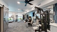 Located at 1888 Brittmoore Rd in Houston, Texas, this fitness center offers a thoughtfully designed workout space across a single floor. The room features a clean, modern aesthetic with white walls, recessed lighting, and ceiling fans that promote airflow and comfort during exercise. Patterned carpet tiles cover the floor throughout, adding both visual interest and durability underfoot.

The cardio section is well-appointed with multiple treadmills, an elliptical trainer, and a rowing machine, all positioned near large windows that allow natural light to fill the space and provide views of the surrounding greenery. A dedicated strength training area includes a multi-functional cable machine system, a dumbbell rack with a full range of free weights, and an adjustable workout bench, supported by a floor-to-ceiling mirror wall that enhances the sense of openness and allows for form monitoring. Stability balls are also available for functional training.

A large flat-screen display is mounted on the mirror wall, and roller shades on the windows offer adjustable light control. The space is accessed via a stylish sliding barn door, adding a contemporary architectural touch. The overall ambiance is bright, clean, and functional, making it a practical and inviting environment for a full-body workout.