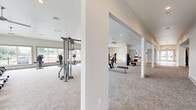 This expansive fitness facility is thoughtfully designed to accommodate a wide range of workout styles and fitness goals. Spread across a single floor, the space is divided into two distinct exercise rooms, each offering a unique training environment. The main exercise area is generously proportioned, featuring plush carpeted flooring, recessed lighting, and multiple ceiling fans that maintain comfortable airflow throughout. Large windows line the perimeter, flooding the space with natural light and offering outdoor views that enhance the overall atmosphere. This primary zone is outfitted with an extensive array of True-brand strength machines, cable systems, treadmills, and cardio equipment, providing comprehensive options for both strength and cardiovascular training. Structural columns subtly define zones within the open layout without disrupting the sense of spaciousness. A secondary exercise room offers a more focused training environment, housing a power rack with free weights, adjustable benches, a mirrored wall, and a dedicated area with dumbbell racks and exercise mats. Additional highlights include a TRX suspension training rig, spin bikes, medicine balls, resistance bands, and a battle rope station. The facility's neutral color palette, white walls, and clean finishes create a bright, motivating atmosphere throughout.